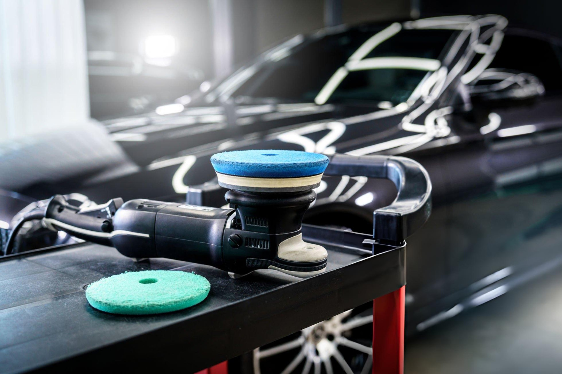 Dual-action car polisher being used on black car hood showing mirror-like polished finish in professional detailing garage